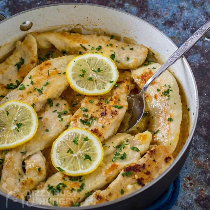 Close up shot of creamy lemon chicken in a skillet garnished with lemon slices and herbs with a spoon.