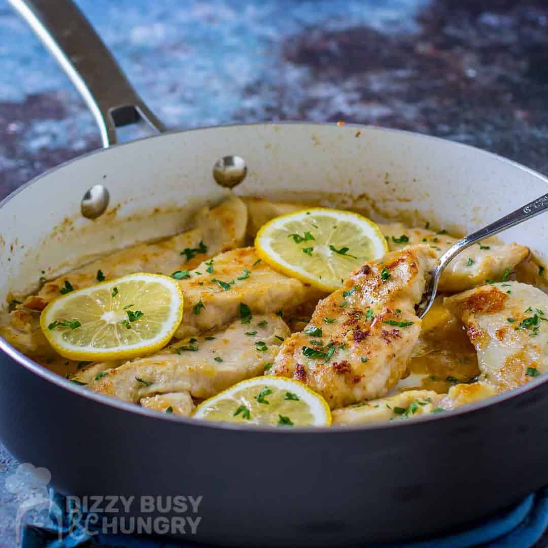Side view of creamy lemon chicken in a skillet garnished with lemon slices and herbs with a spoon.
