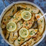 Overhead shot of creamy lemon chicken garnished with herbs and lemon slices in a skillet with a spoon.