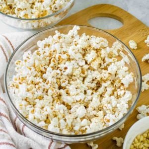 Close up shot of popcorn with homemade seasoning in a clear bowl on a wooden cutting board with popcorn sprinkled around.