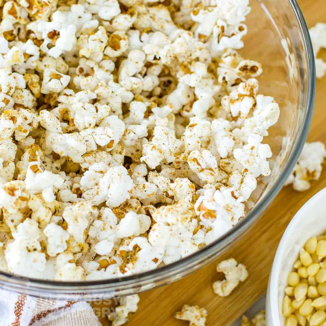 Close up overhead shot of homemade popcorn seasoning in a clear bowl on a wooden cutting board with popcorn sprinkled around.