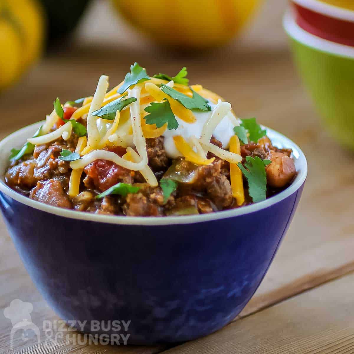 Close up shot of chili with salsa in a small blue bowl with bowls stacked on the side and colorful pumpkins in the background.