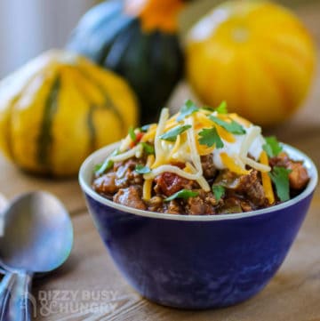 Side view of chili with salsa in a small blue bowl with a spoon on the side and colorful pumpkins in the background.