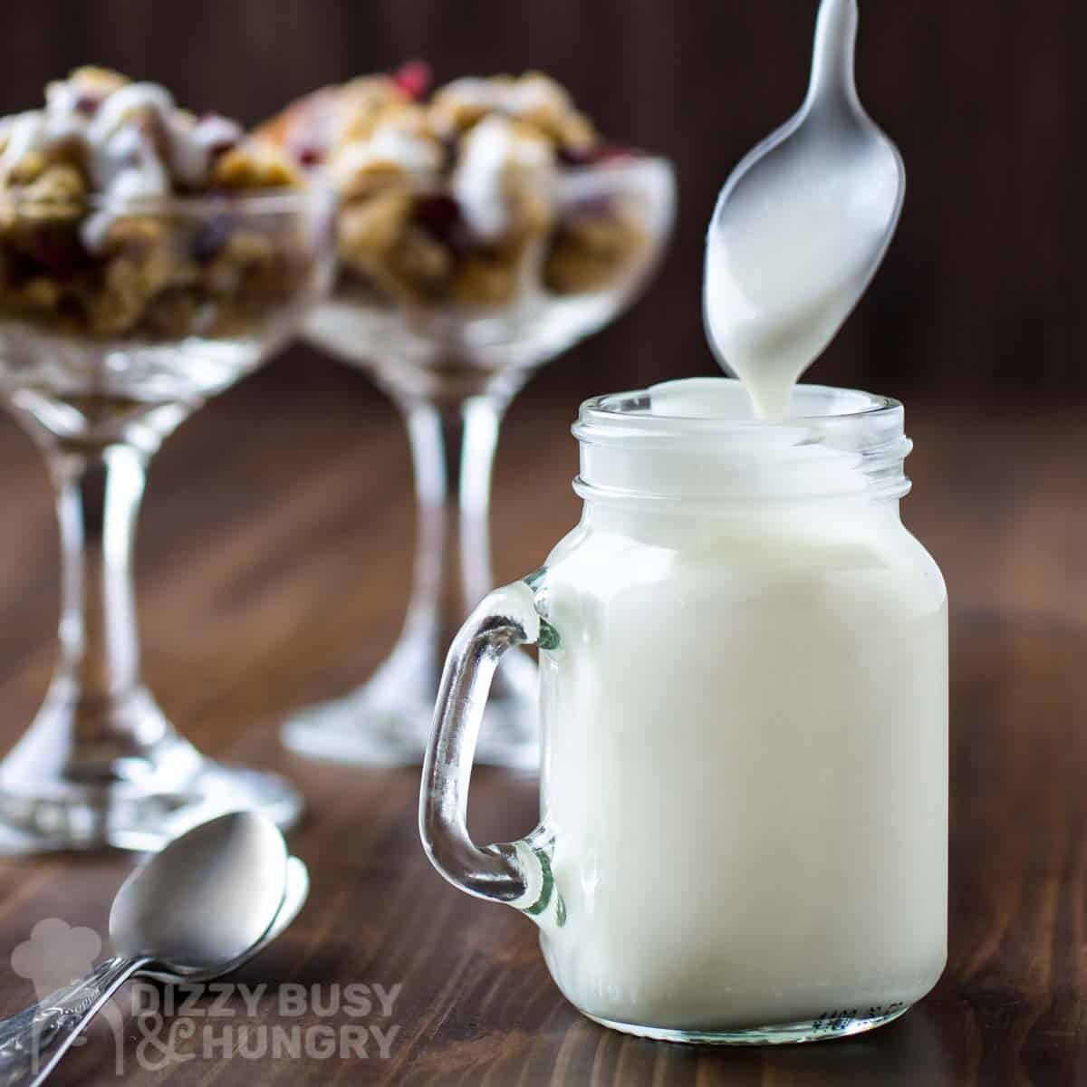 Side view of yogurt icing being drizzled into a mason jar from a spoon with glasses and spoons in the background.