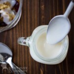 Overhead shot of a spoon dipping into a mason jar with greek yogurt icing on a wooden surface.