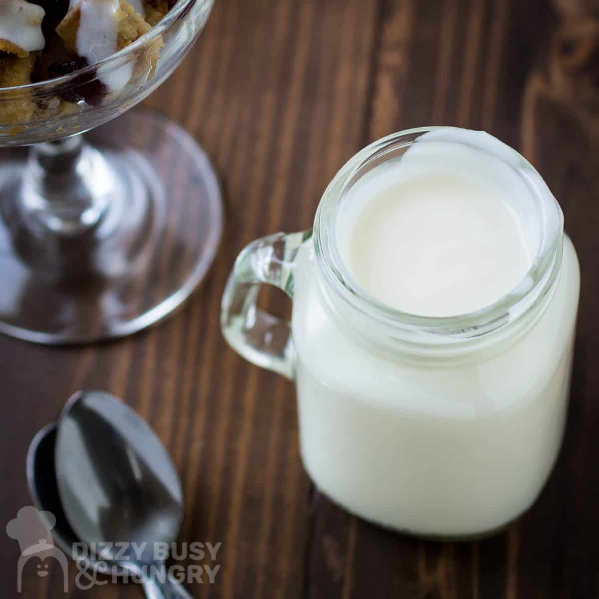 Side view of greek yogurt in a mason jar with a glass and spoons on the side on a wooden surface.