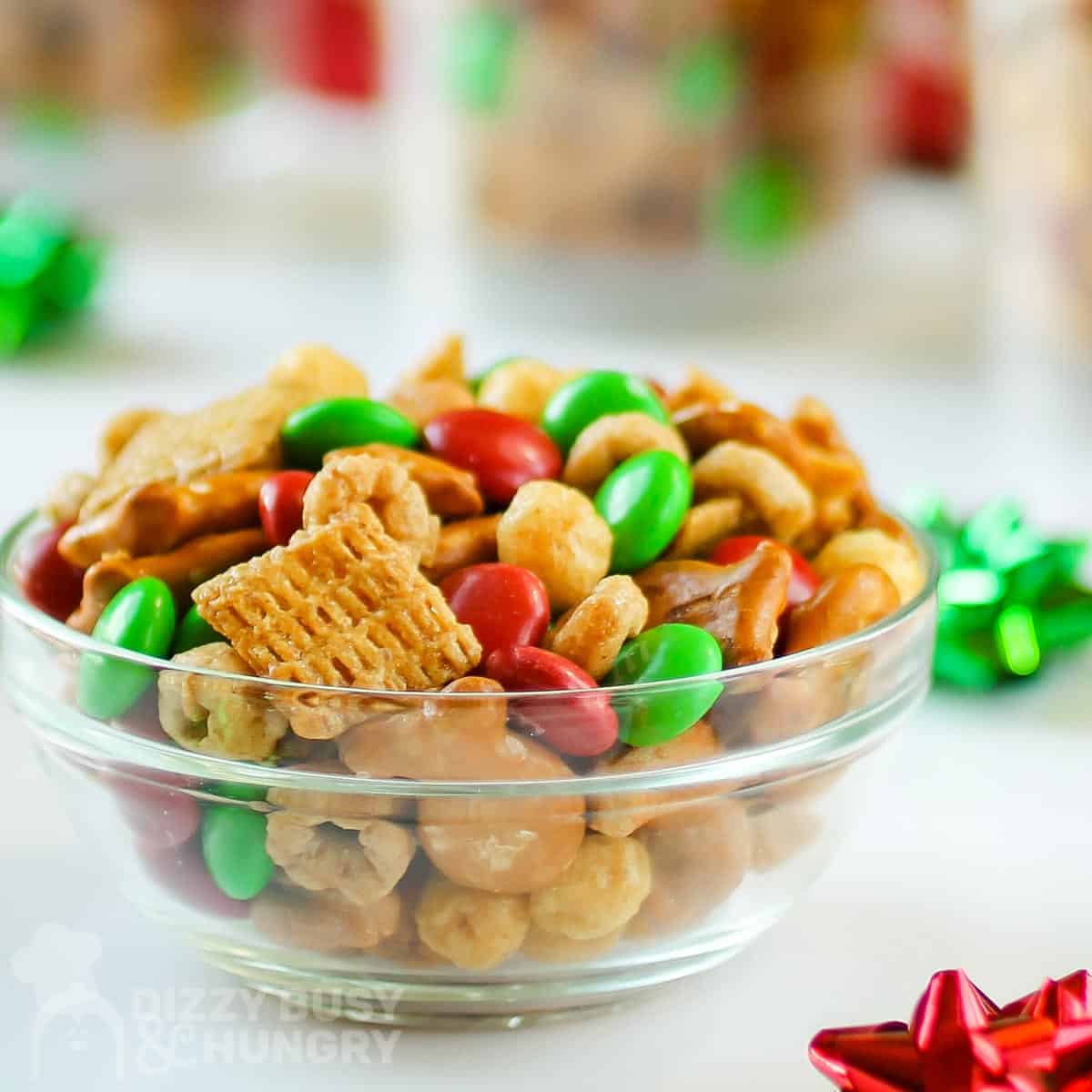Side view of Christmas snack mix in a white bowl with gift bows scattered around on a white table, with more jars of he snack mix in the background.