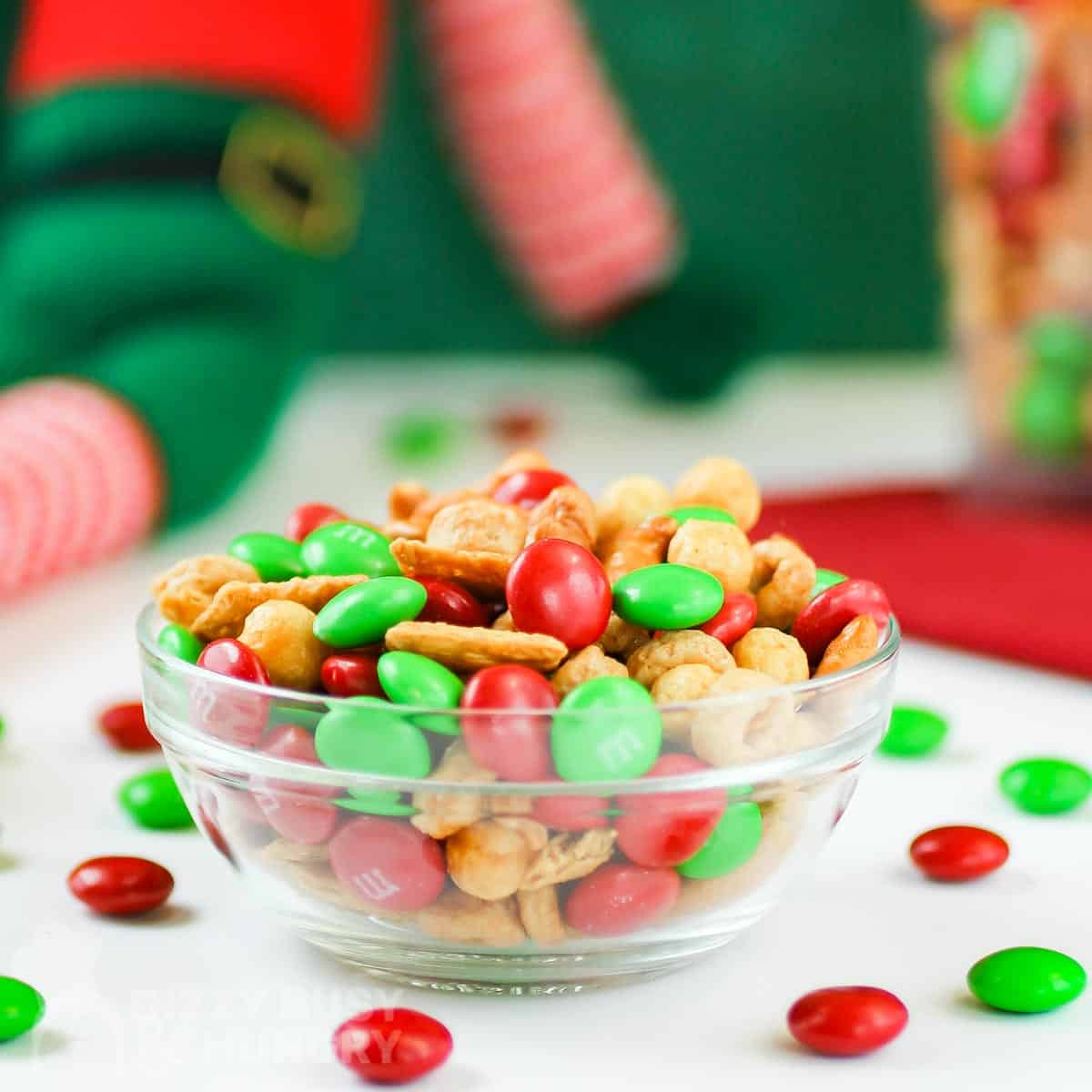 Side view of Christmas snack mix in a white bowl with M&Ms scattered around on a white table, with Christmas decorations in the background.