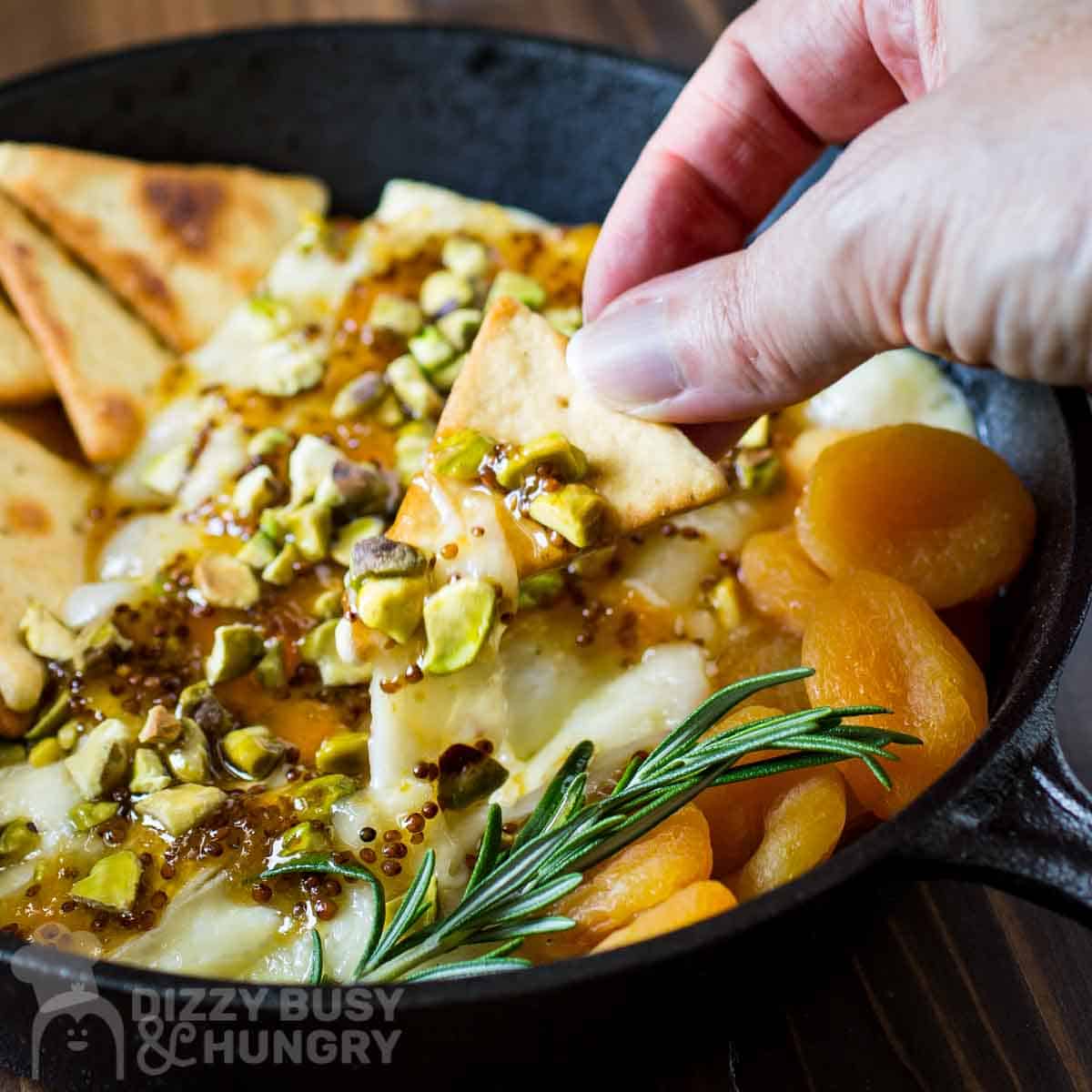 Close up shot of a pita cracker being dipped into baked brie with more crackers, apricots, and herbs on the side in a skillet on a wooden surface.
