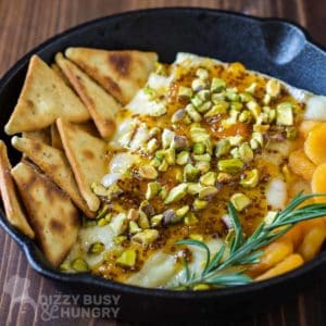 Close up shot of baked brie in a skillet with pita chips, apricots and herbs surrounding it on a wooden surface.