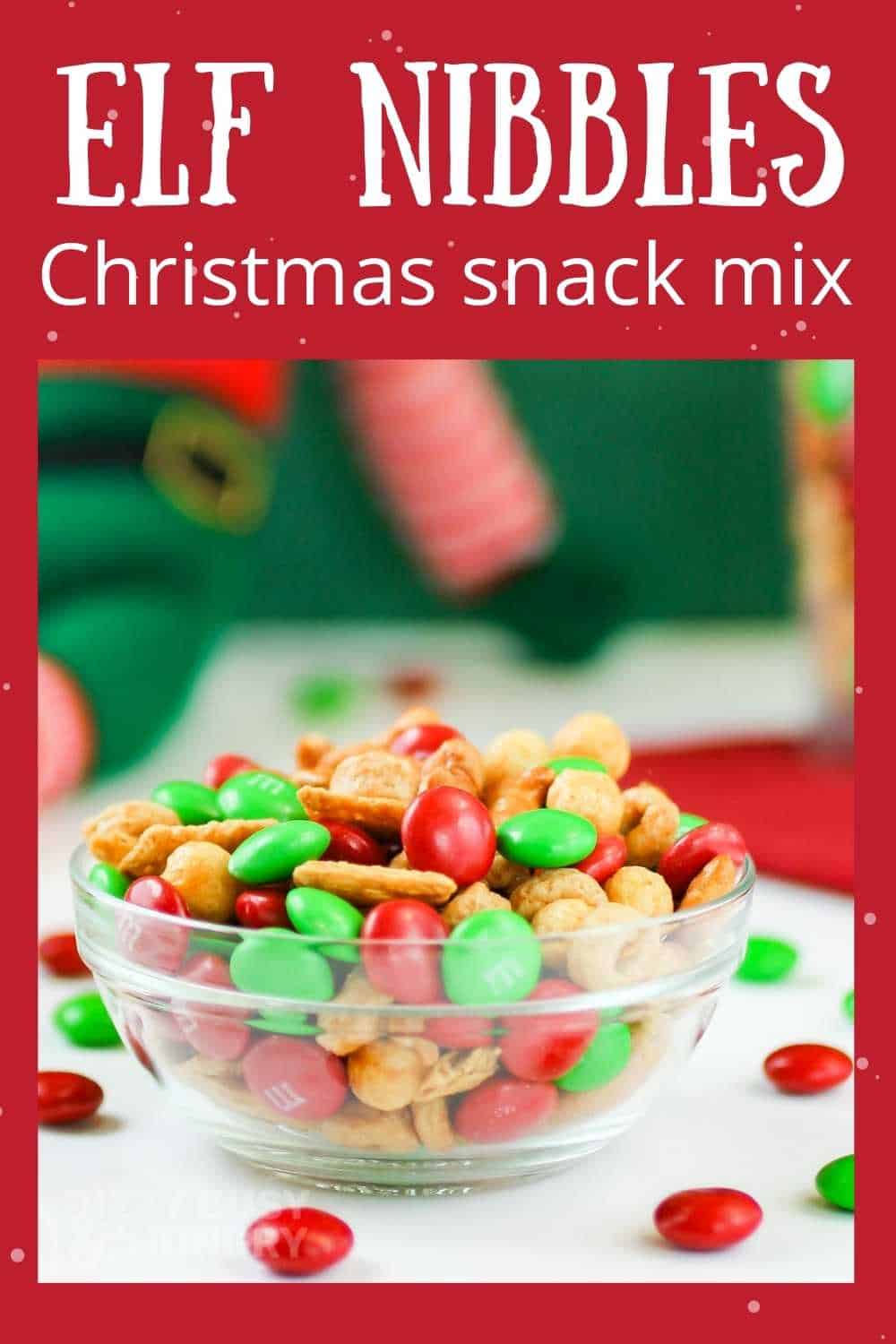 Side view of Christmas snack mix in a white bowl with M&Ms scattered around on a white table, with Christmas decorations in the background.