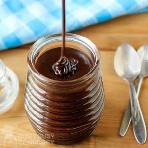 Side view of chocolate sauce being drizzled into a clear glass with a blue plaid cloth and spoons in the background.