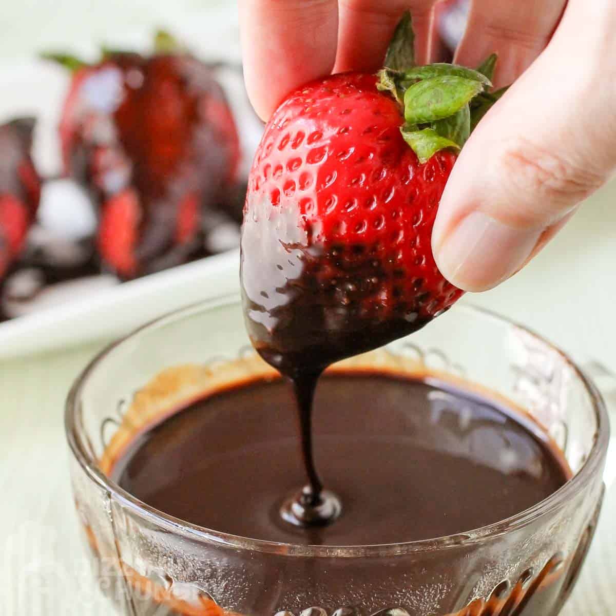 Close up shot of a strawberry being dipped into a clear bowl of chocolate sauce with more chocolate covered strawberries in the background.