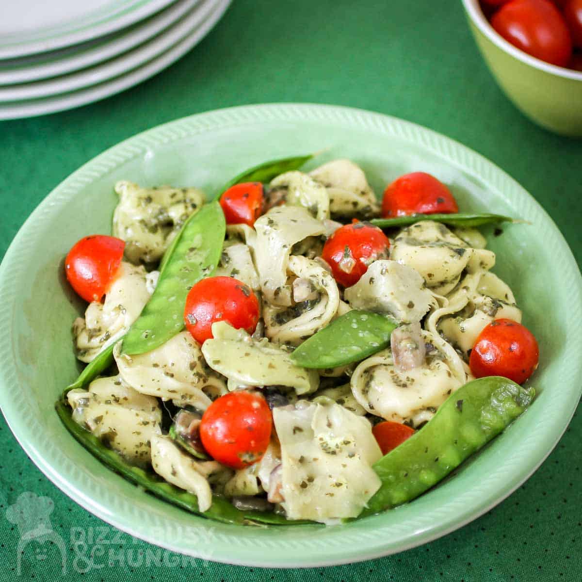 Overhead shot of pesto tortellini in a green bowl with a bowl of grape tomatoes and plates in the background.