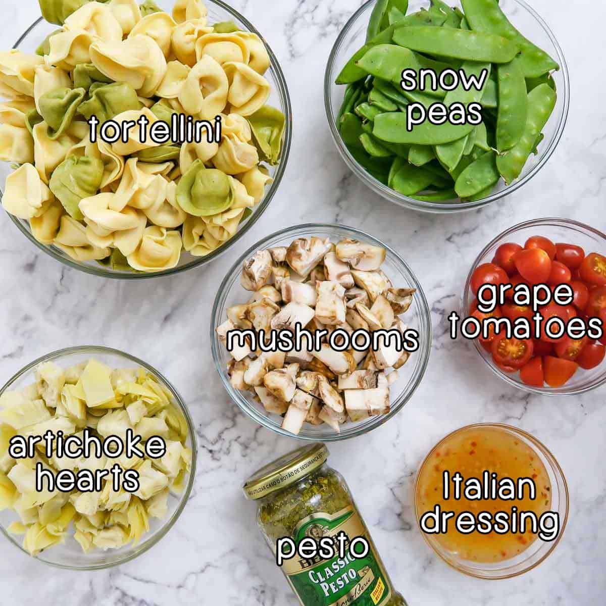 Overhead shot of ingredients- tortellini, snow peas, mushrooms, grape tomatoes, artichoke hearts, pesto, and Italian dressing.