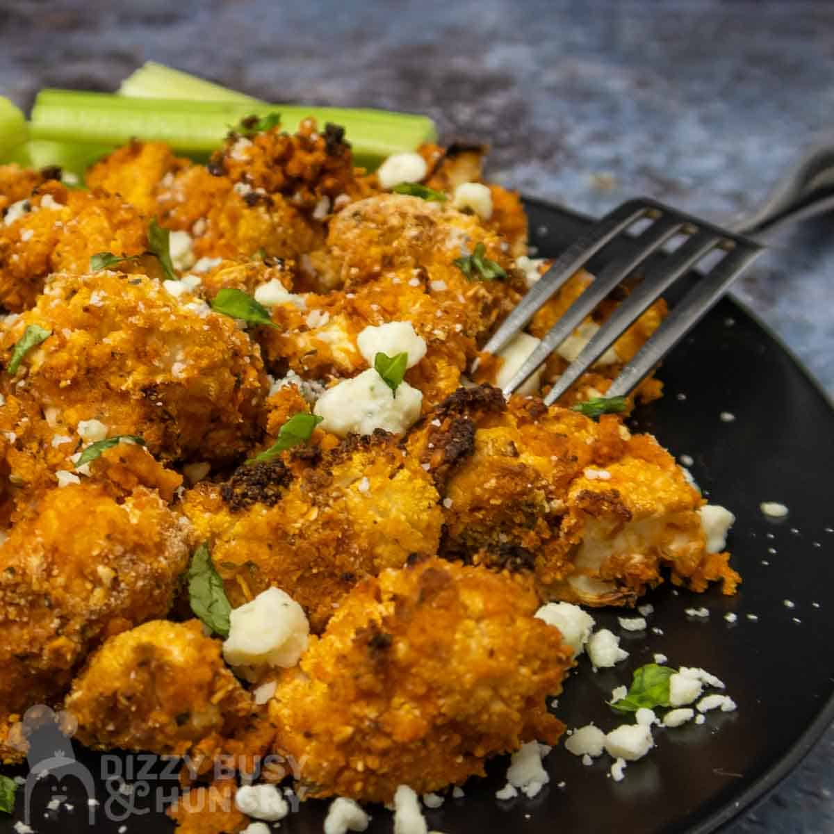 Close up shot of buffalo cauliflower on a black plate sprinkled with cheese crumbles and herbs with a fork and celery on the side.