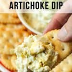 Close up shot of a person holding up a cracker dipped in the jalapeno artichoke dip with the bowl of dip and crackers in the background.