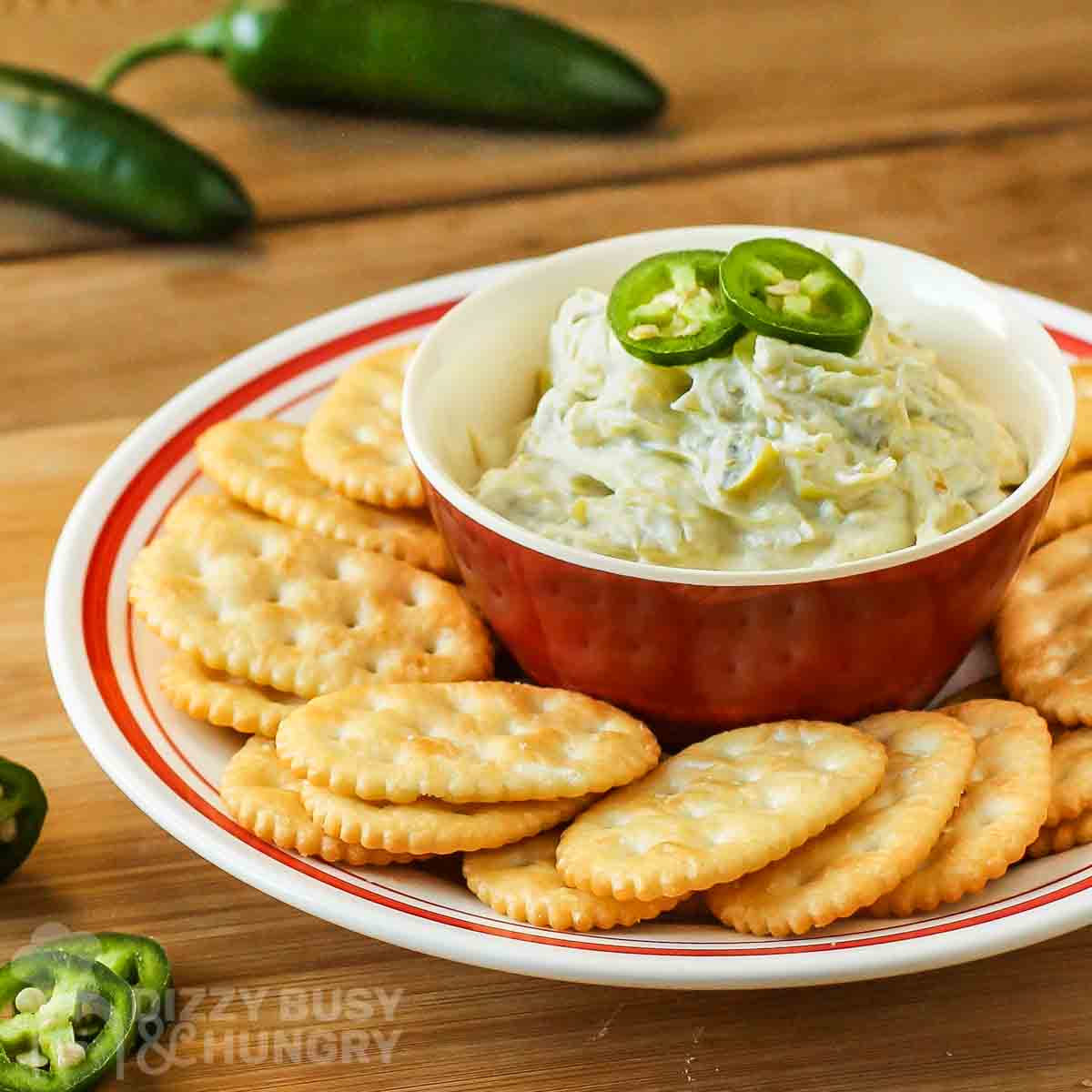 Side view of jalapeno artichoke dip in a red bowl surrounded by a ring of crackers on a red and white plate.