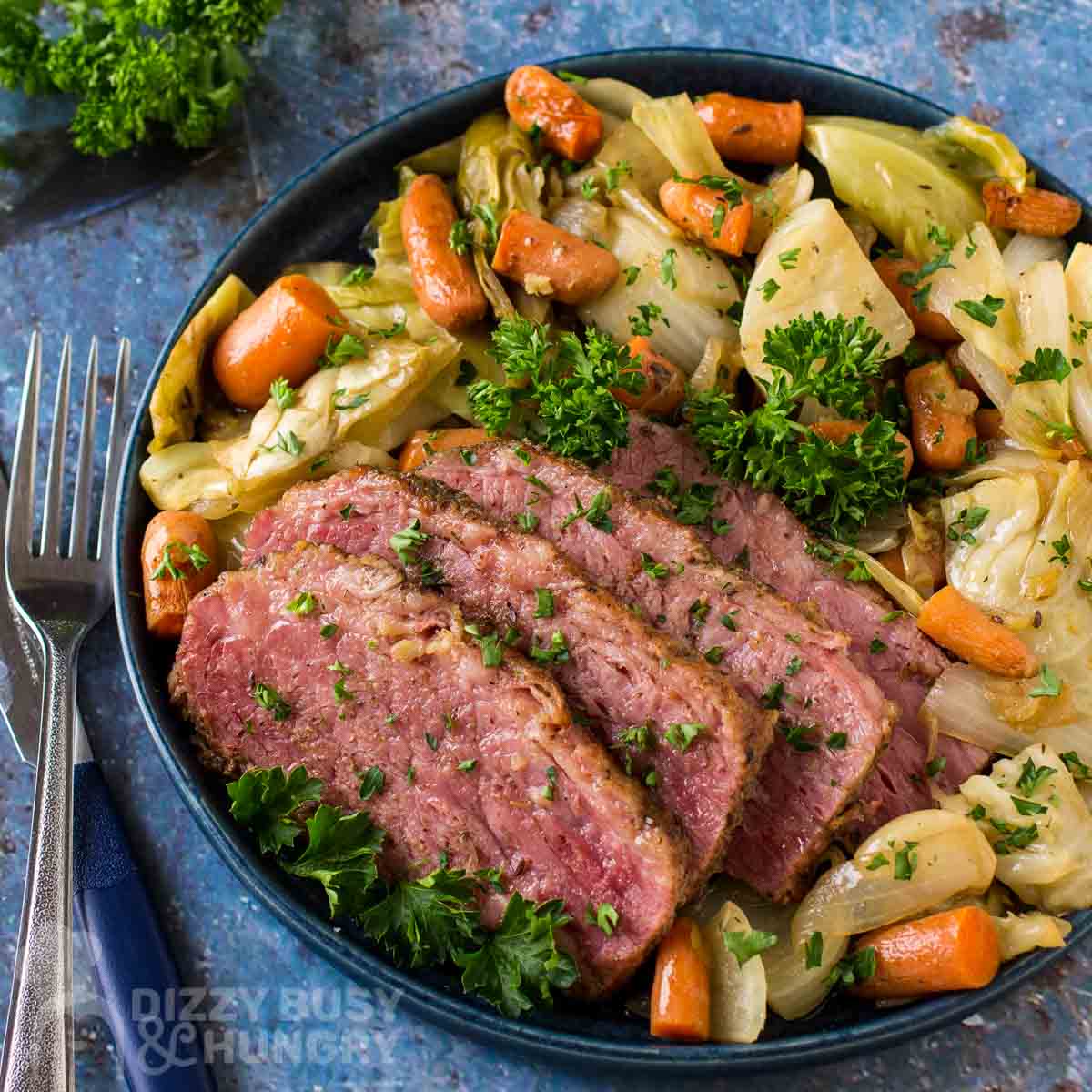 Overhead shot of braised corned beef with vegetables garnished with herbs on a black plate with forks and herbs on the side on a blue cloth.