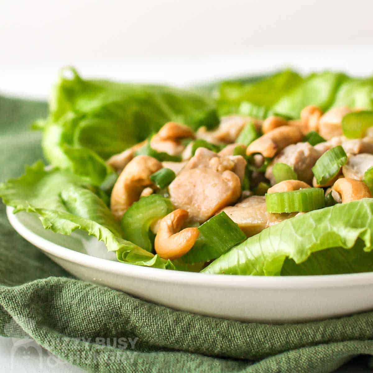 Side shot of lettuce wrap garnished with cashews and scallions on a white plate on top of an olive green napkin.