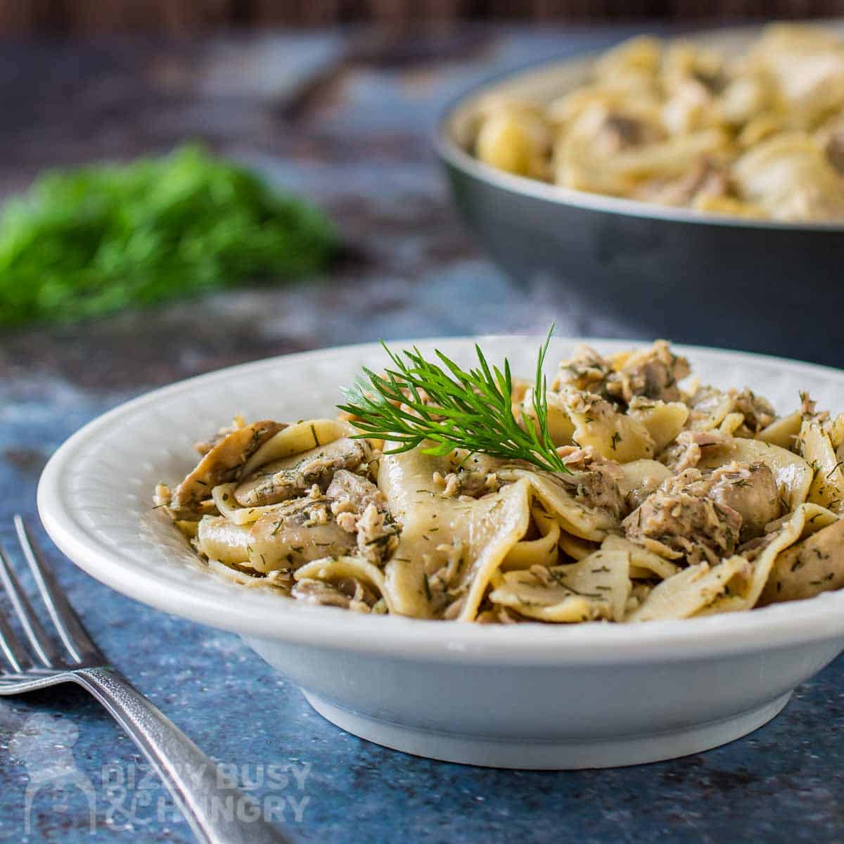 Side close up shot of lemon dill tuna and noodles garnished with herbs in a white bowl on a blue placemat with silverware on the side.