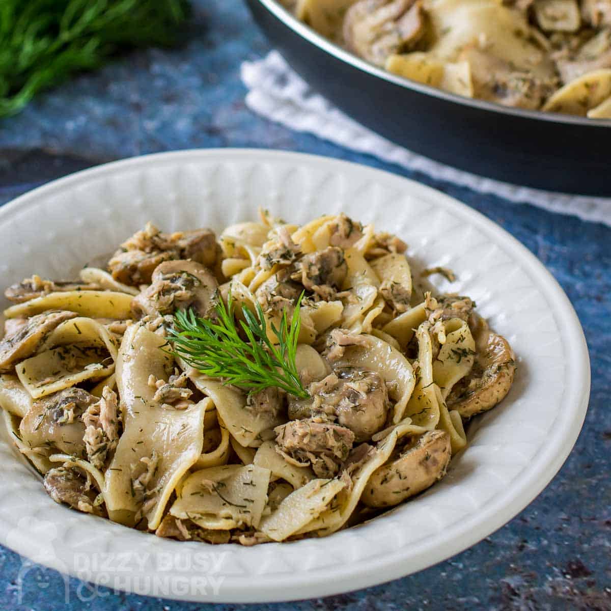 Side view of lemon dill tuna and noodles in a white bowl garnished with herbs on a blue place setting with more herbs in the background.