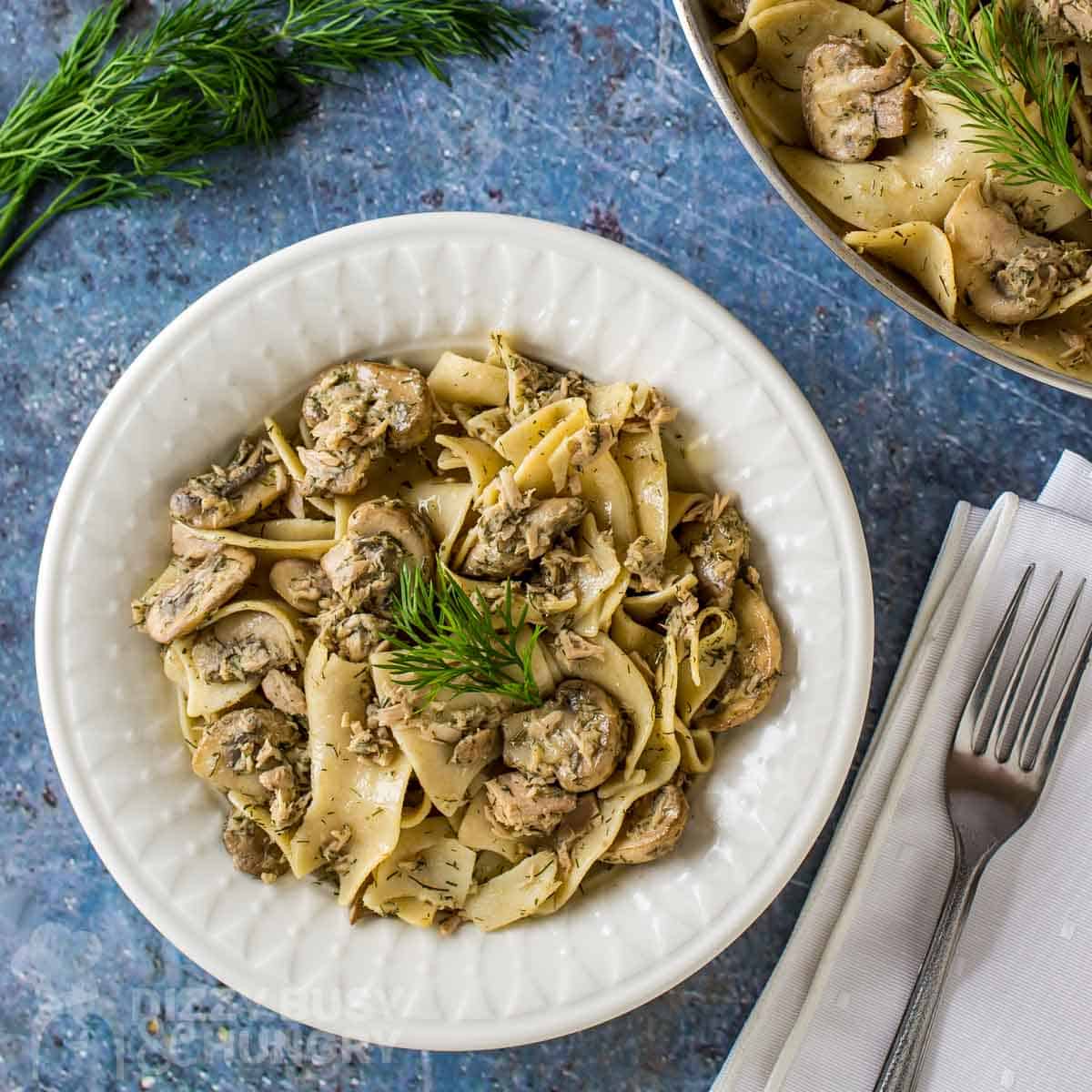Overhead shot of lemon dill tuna and noodles garnished with herbs in a white bowl on a blue placemat with herbs and silverware on the side.
