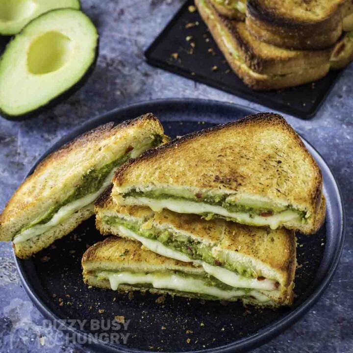 Side shot of multiple triangle halves of avocado grilled cheese on a black plate with more on a black plate and an avocado in the background.