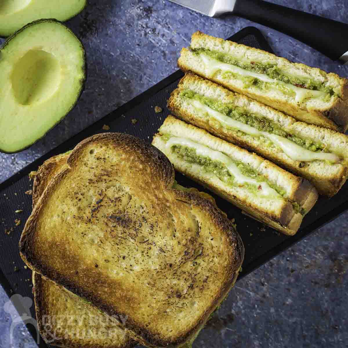 Overhead shot of full and halved avocado grilled cheese on a black rectangle plate on a blue marbled surface with a knife and halved avocados on the side.