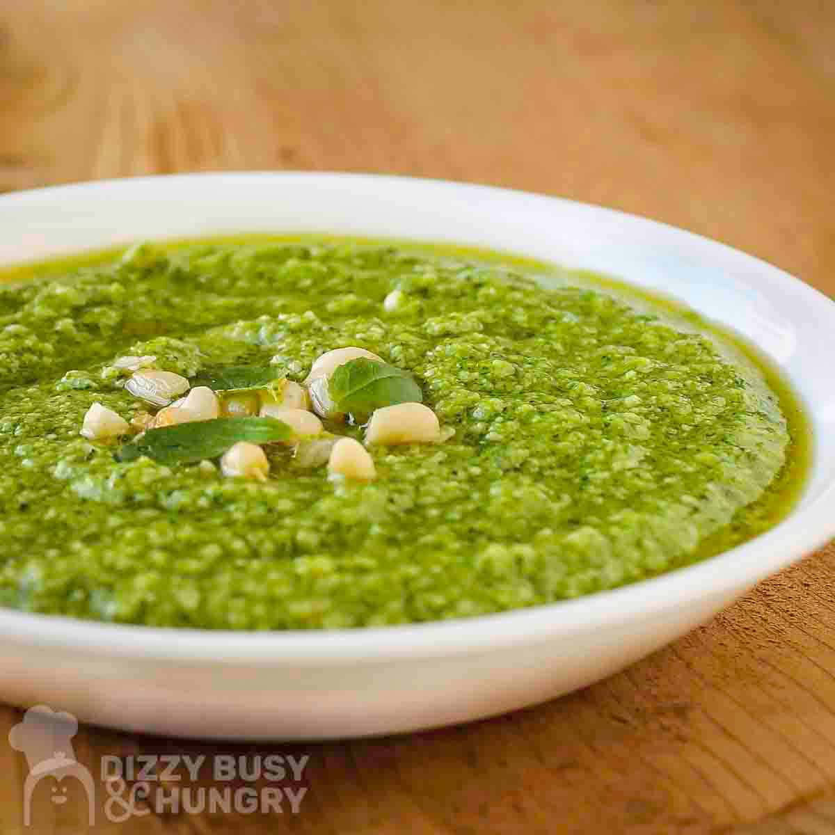 Side close up view of broccoli pesto in a white bowl garnished with basil and pine nuts with more basil on a wooden cutting board in the background.