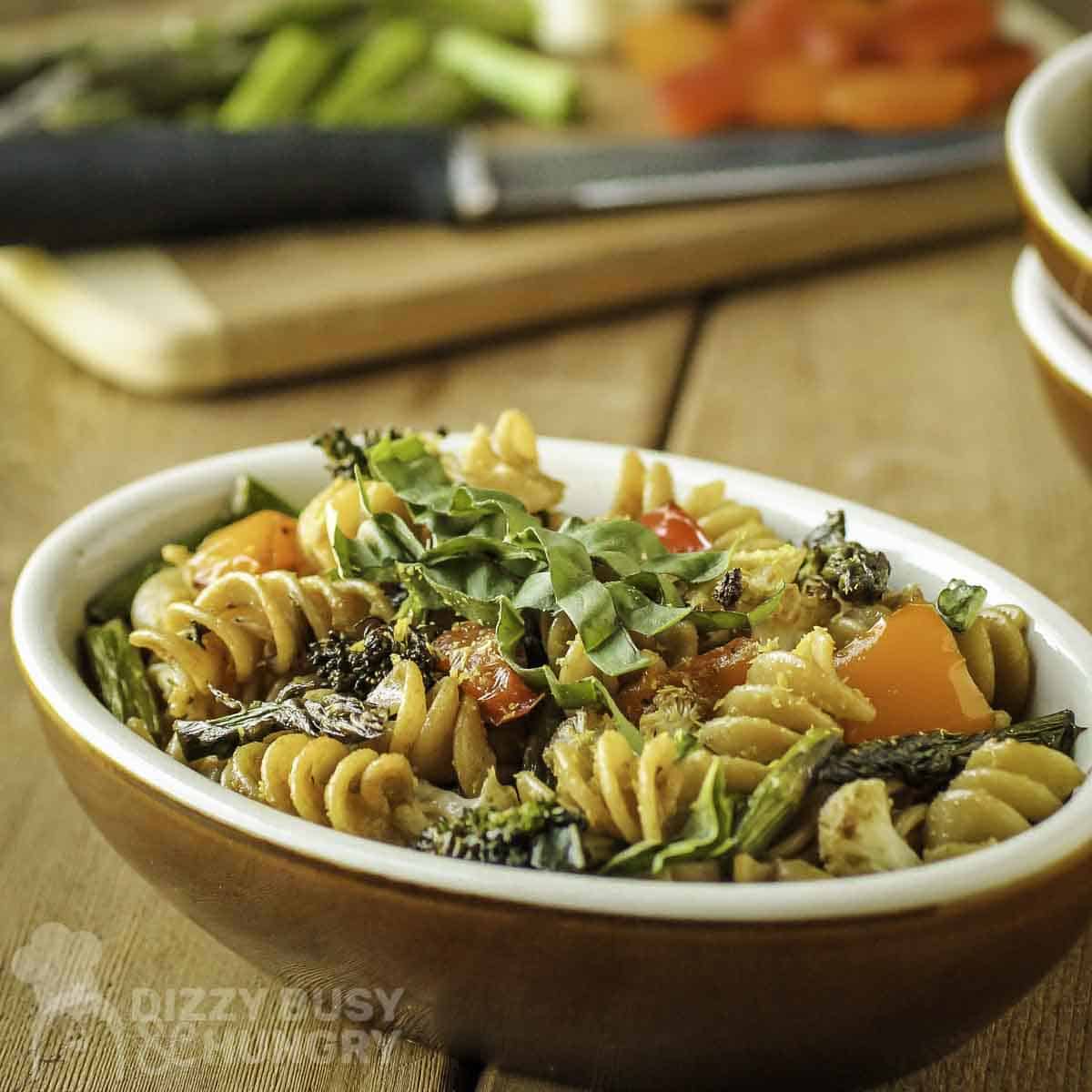 Side shot of veggie rotini in an orange and white oval bowl with chopped veggies on a wooden cutting board in the background.