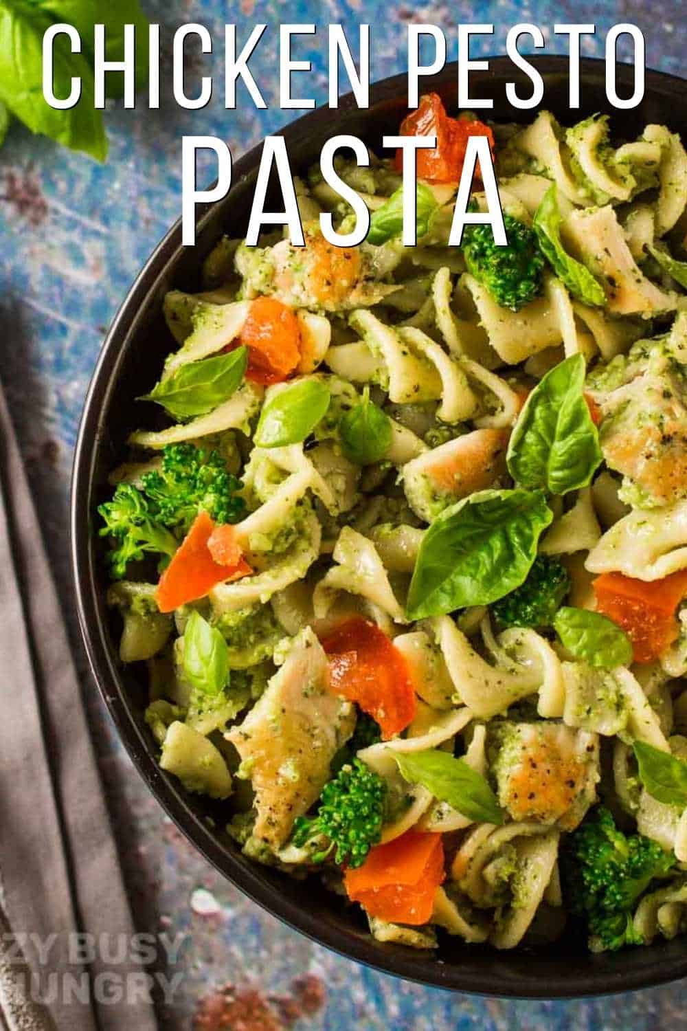 Overhead shot of chicken pesto pasta garnished with basil in a black bowl on a blue surface with a napkin on the side.