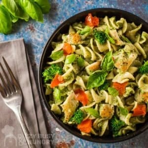 Overhead shot of chicken pesto pasta garnished with basil in a black bowl on a blue surface with a fork and more basil on the side.