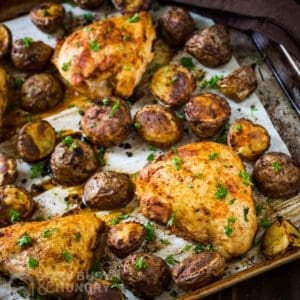 Side shot of chicken tray bake on a baking sheet lined with parchment paper garnished with herbs.