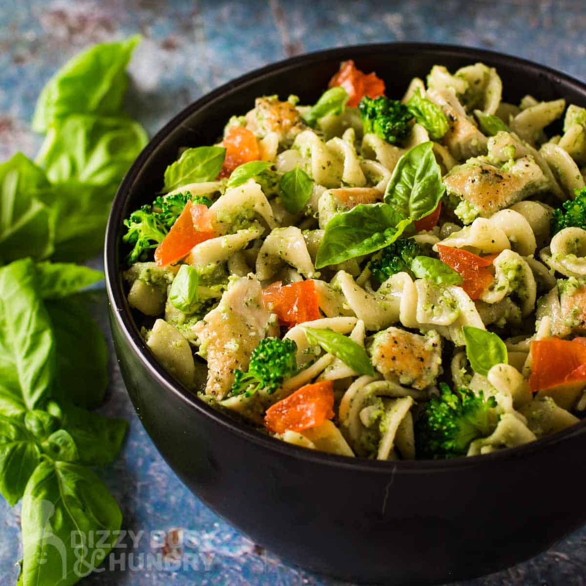 Side shot of chicken pesto pasta garnished with basil in a black bowl on a blue surface with more basil on the side.