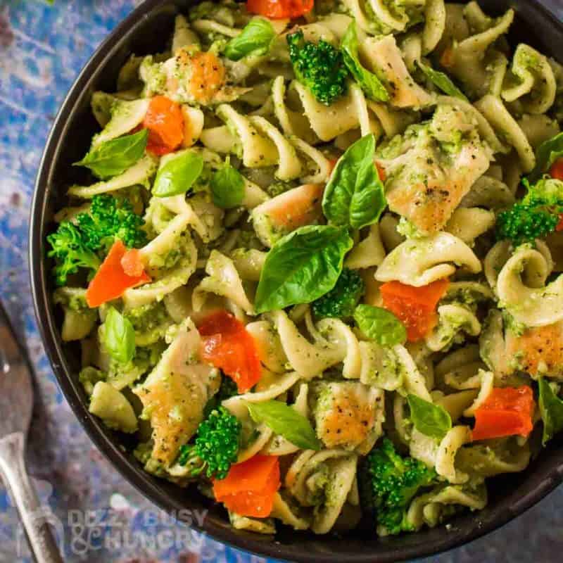 Overhead close up shot of chicken pesto pasta garnished with basil in a black bowl on a blue surface.