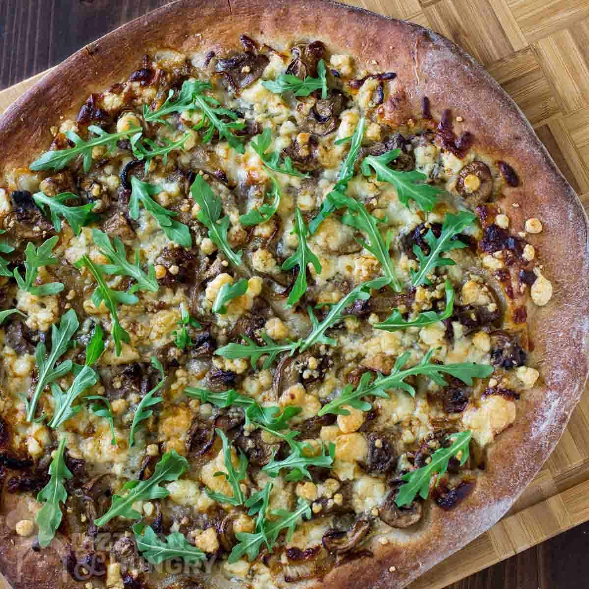 Overhead shot of whole steak pizza garnished with arugula on a wooden cutting board.