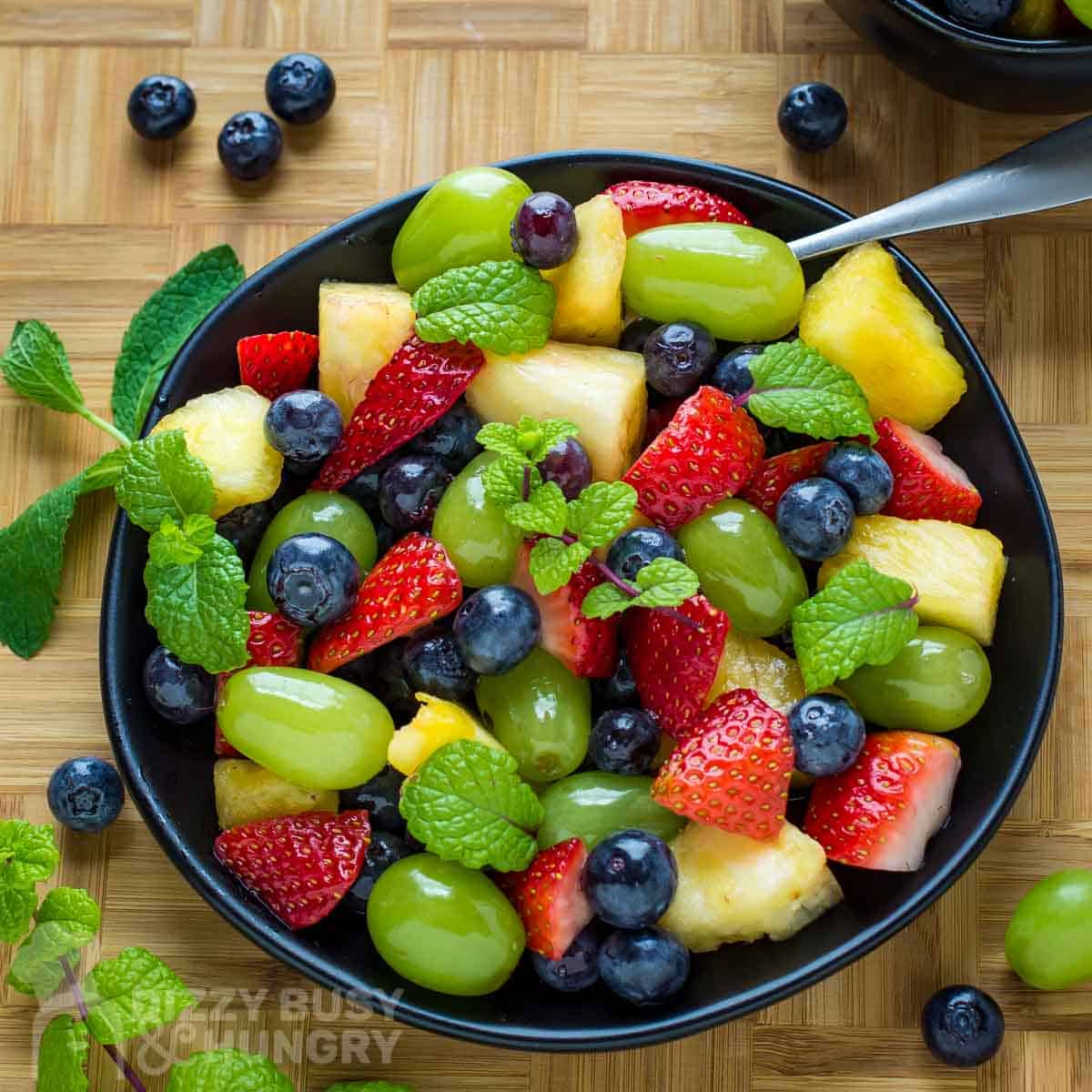 Overhead shot of berry fruit salad garnished with mint on a black plate with a spoon and more mint on the side.