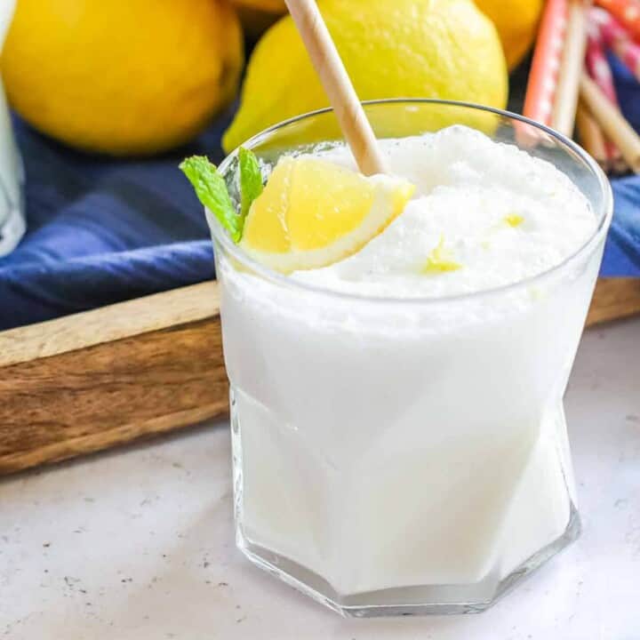 Side shot of frosted lemonade in a clear glass with two straws and a lemon and mint as a garnish with a blue cloth and lemons in the background.