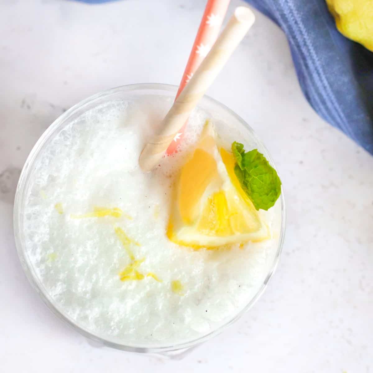 Overhead close up shot of frosted lemonade garnished with lemon and mint with two straws and a blue cloth on the side.