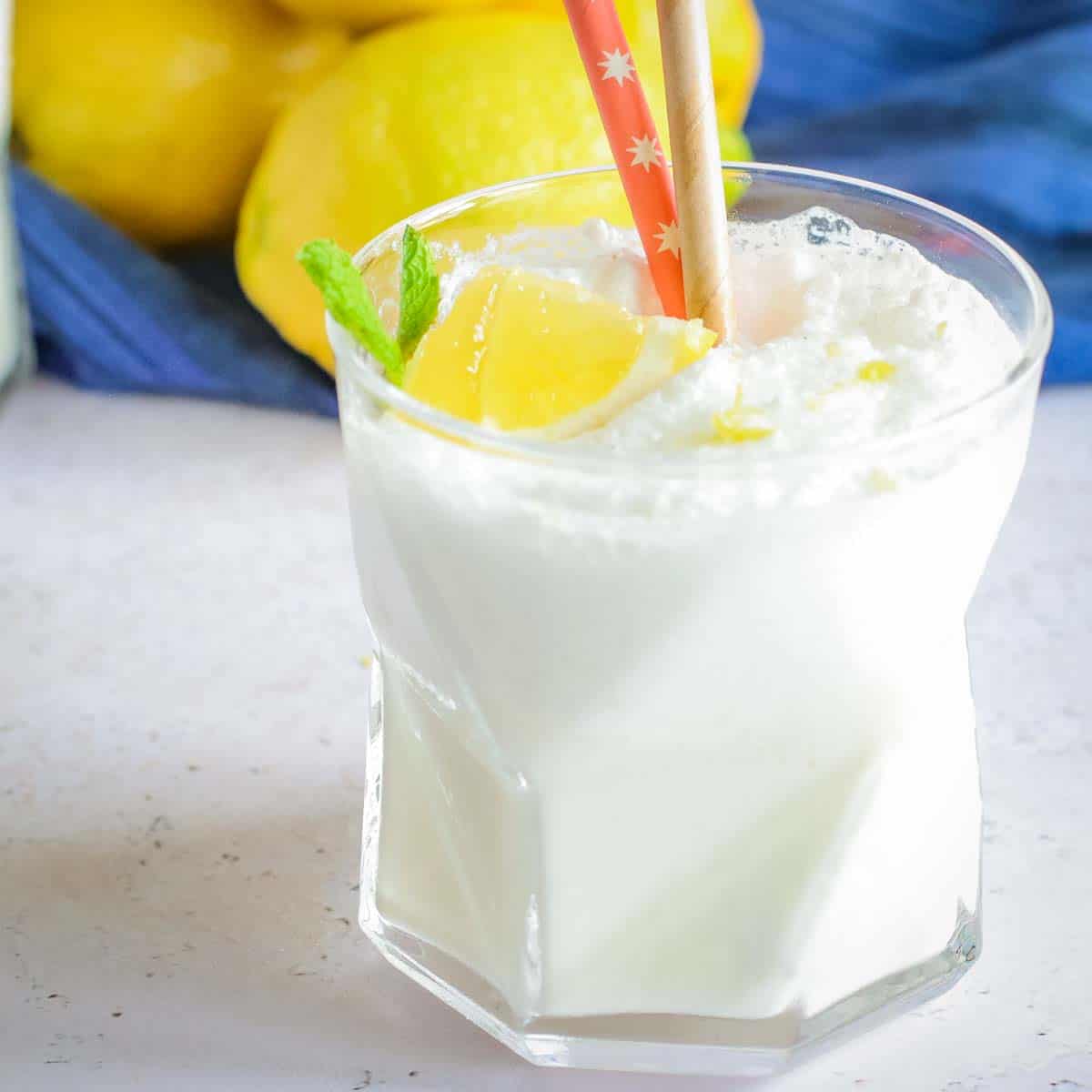 Close up side shot of a chick fil a frozen lemonade in a clear glass with two straws and a lemon and mint as a garnish with a blue cloth and lemons in the background.