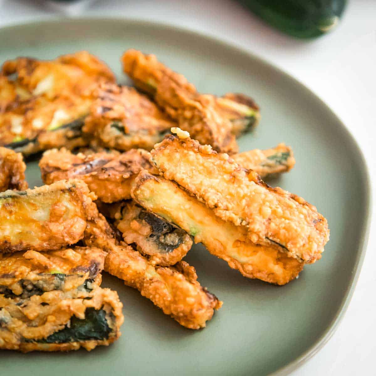Side shot of multiple zucchini fries on a light green plate with a white background and a whole zucchini in the background.