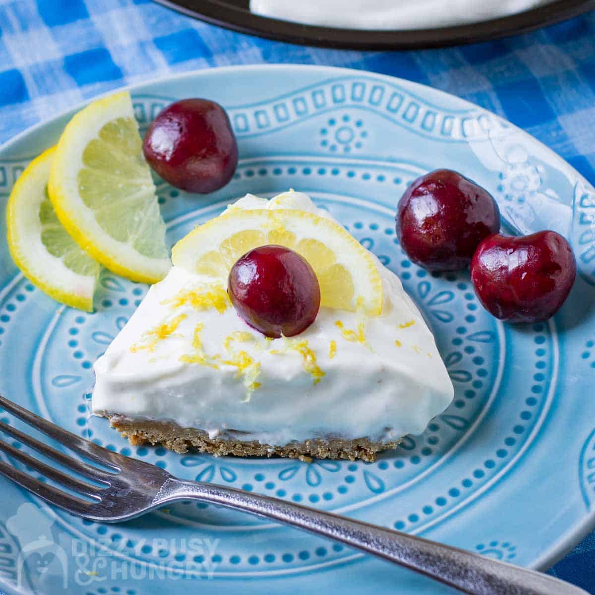 Close up shot of a slice of lemon pie garnished with a whole cherry on a blue plate with a fork, lemon slices, and more cherries on the side.