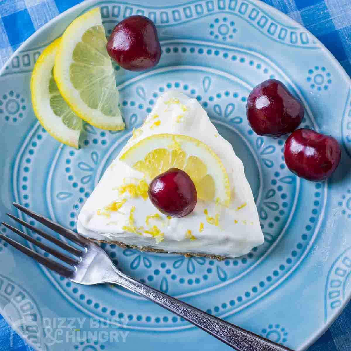 Overhead shot of a slice of lemon pie garnished with a whole cherry on a blue plate with a fork, lemon slices, and more cherries on the side.