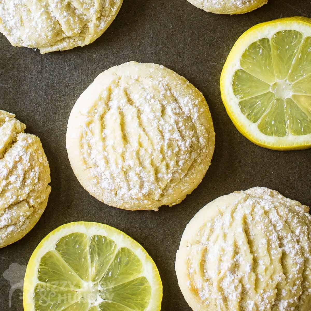 Overhead shot of lemon cookies with powdered sugar on a brown surface with slices of lemons on the side.