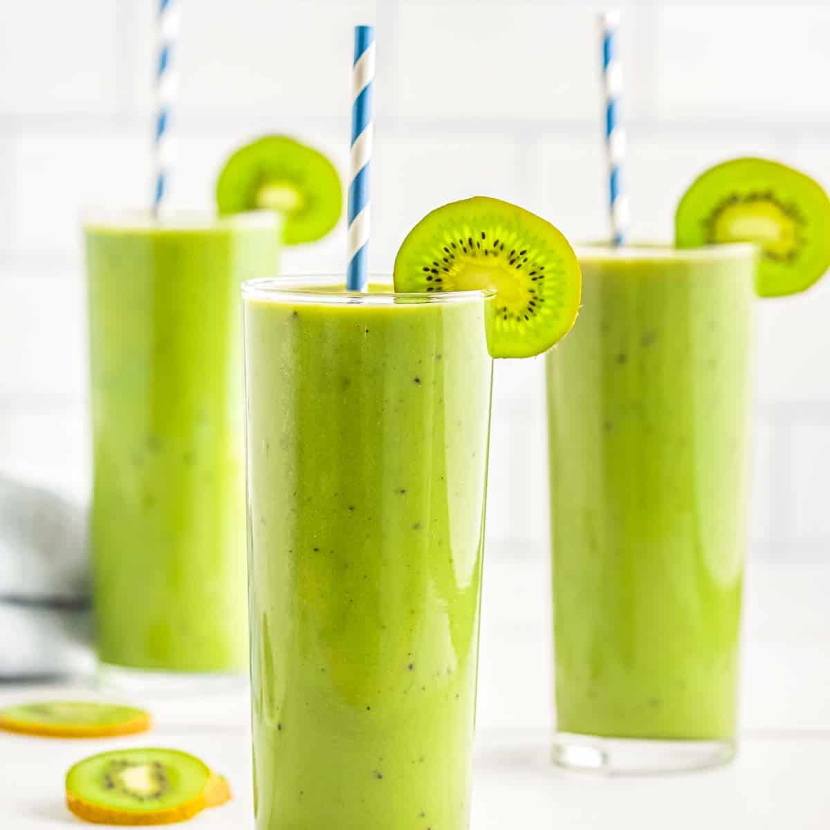Side shot of kiwi banana match smoothie in a tall clear glass with a slice of kiwi and a straw with more glasses in the background.