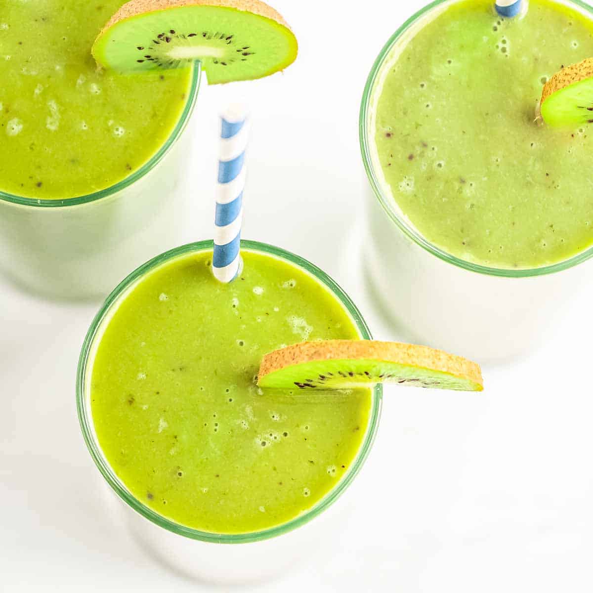 Overhead shot of three glasses of kiwi banana smoothie garnished with a slice of kiwi and a straw on a white surface.