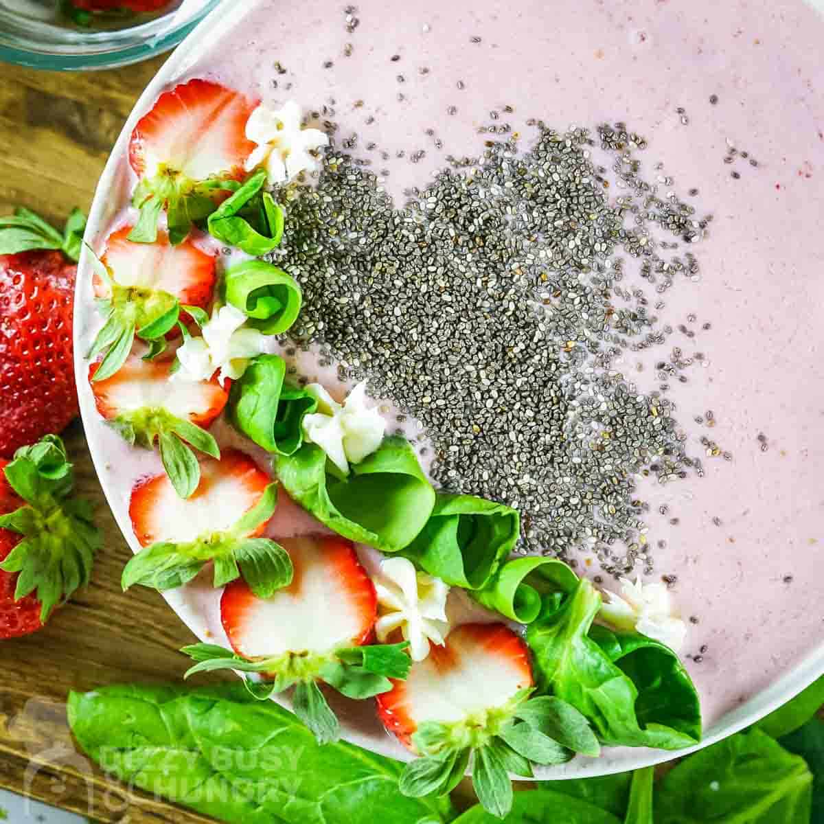 Overhead shot of pink smoothie bowl garnished with flax seeds, sliced strawberries, and mint rolls on a wooden surface with more mint and strawberries on the side.