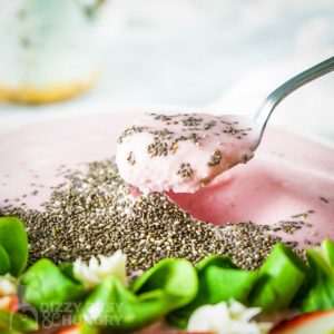 Side close up shot of a spoon in a smoothie bowl garnished with flax seeds, mint, and sliced strawberries on a wooden surface with more mint on the side.