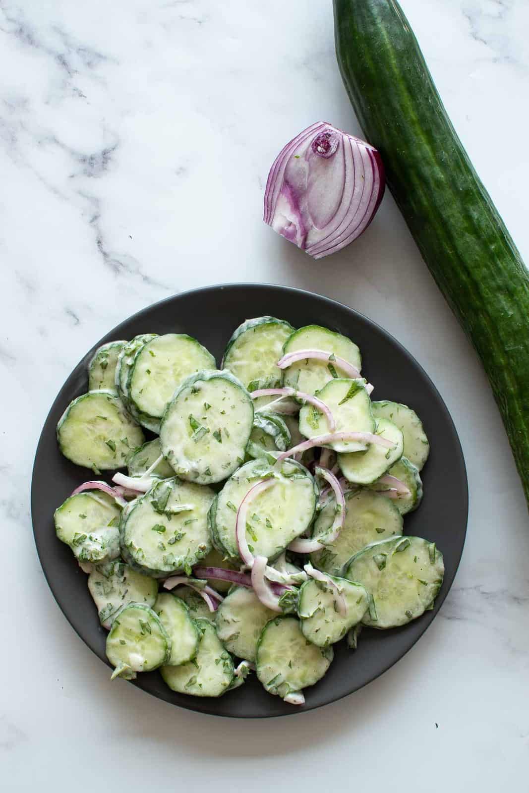 A bowl of cucumber salad viewed from above on a white counter with cucumber and onion on the side.